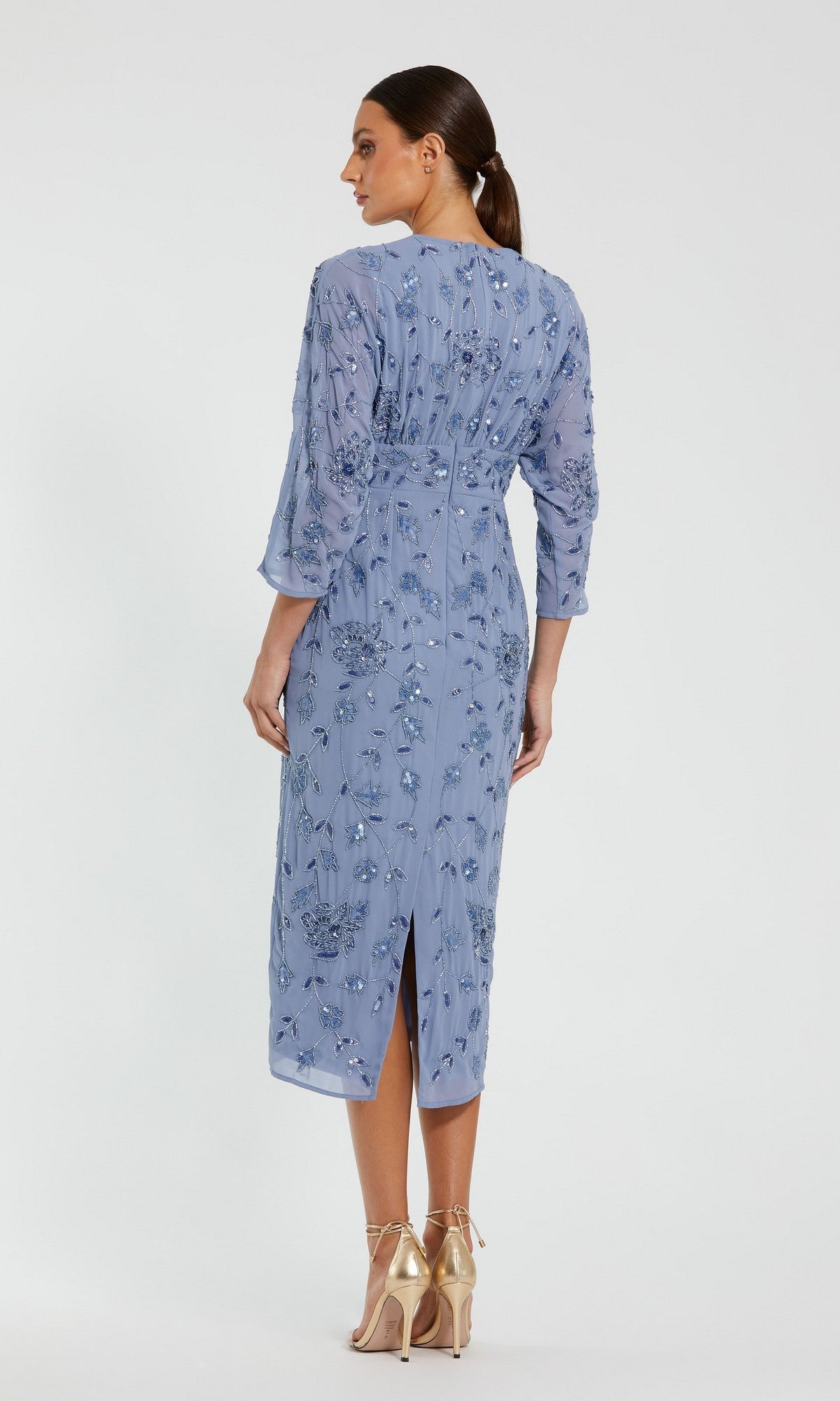 A woman stands with her back to the camera, wearing the Mac Duggal 94141 Midi Party Dress—a light blue, long-sleeve dress featuring intricate embroidery and modest cocktail styling—paired with gold high-heeled shoes against a plain white background.